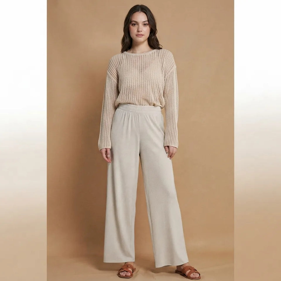 A woman stands against a warm beige studio backdrop wearing a beige ribbed knit long-sleeve top and light beige wide-leg pants with brown sandals.
