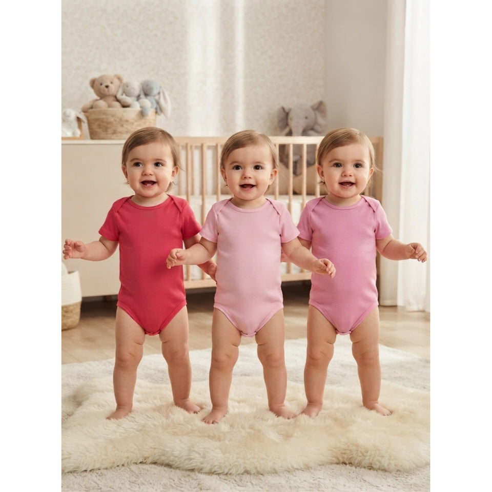 Three babies standing on a fluffy cream rug in a softly lit nursery, each wearing a short-sleeve bodysuit in a different pink shade: a dark raspberry pink, a light bubblegum pink, and a medium pastel pink.