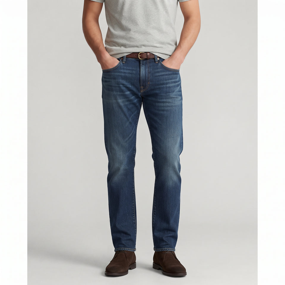 A man wearing a pair of medium-wash blue jeans with a light gray t-shirt and brown suede boots, standing against a light gray studio background.