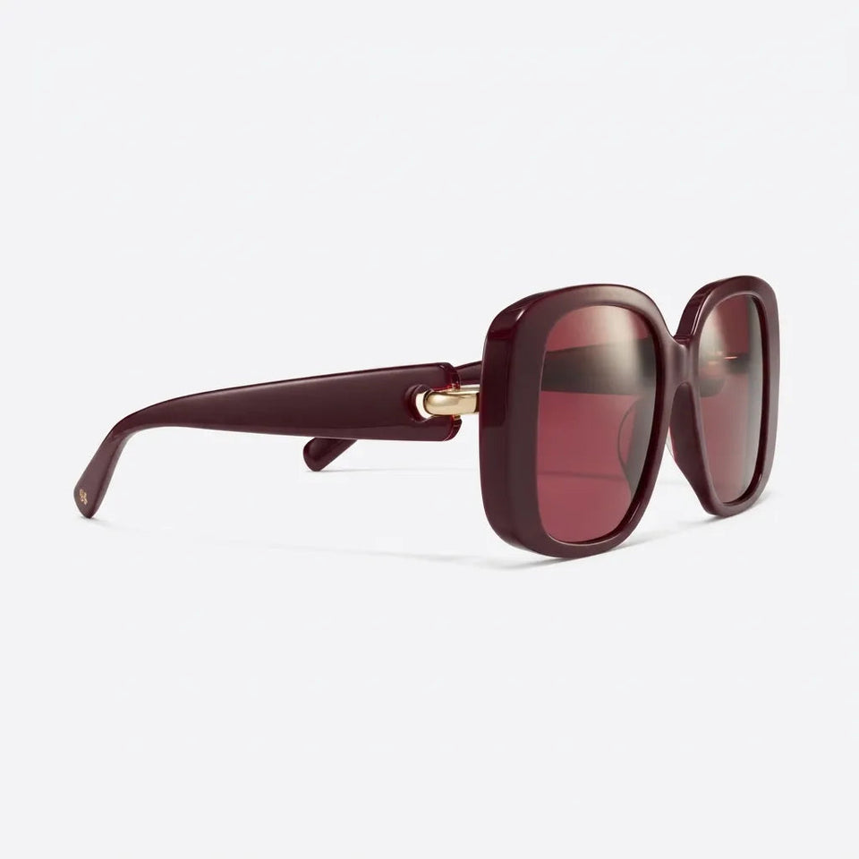 Oversized square sunglasses with a translucent dark burgundy acetate frame and matching tinted lenses, featuring a small gold metal accent at the hinge, shown on a light gray background.