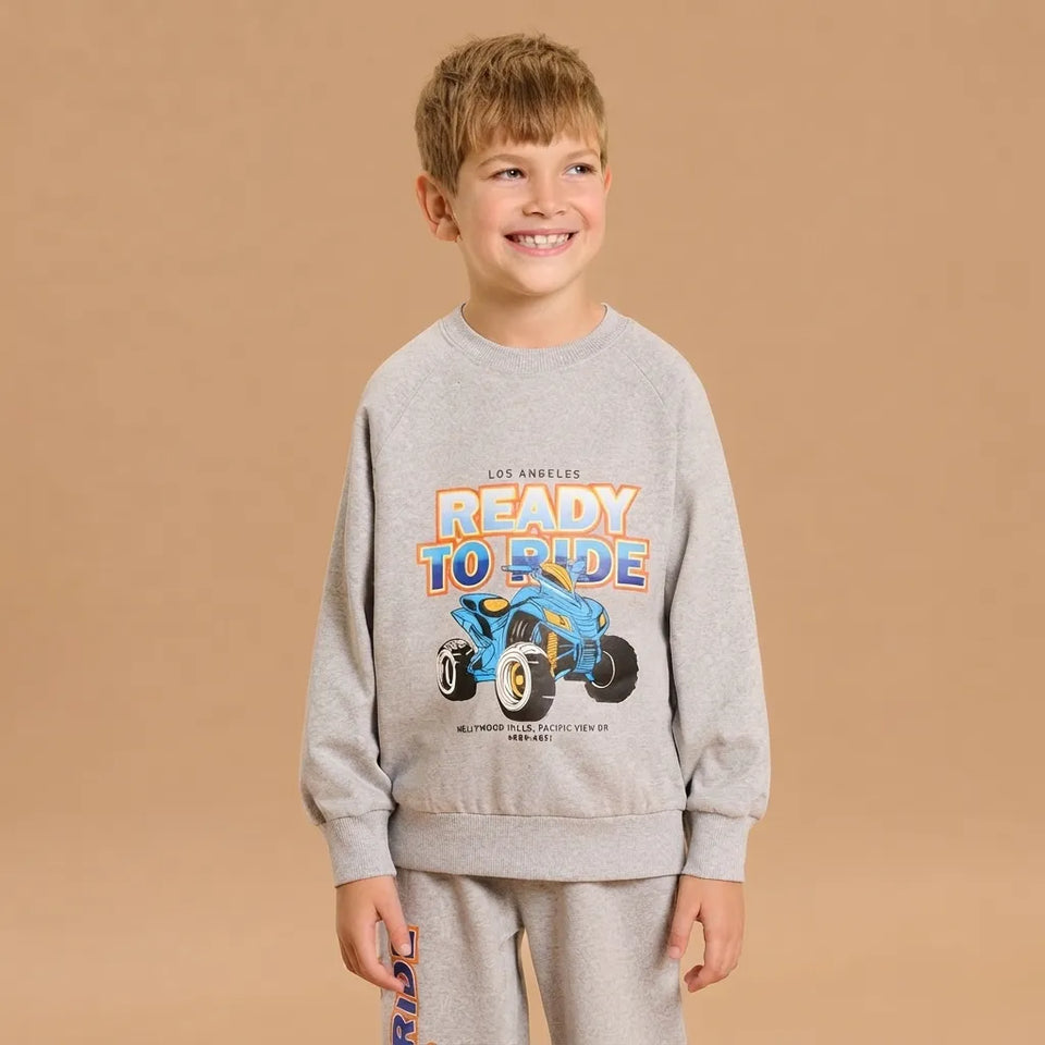 A young boy smiling while wearing a light gray crewneck sweatshirt with a colorful 'Ready to Ride' graphic and matching gray sweatpants, set against a solid tan studio background.