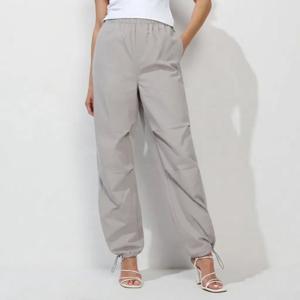 A model wearing light gray wide-leg jogger pants with an elastic waistband and drawstring ankle cuffs, paired with white strappy high-heeled sandals against a white studio background.