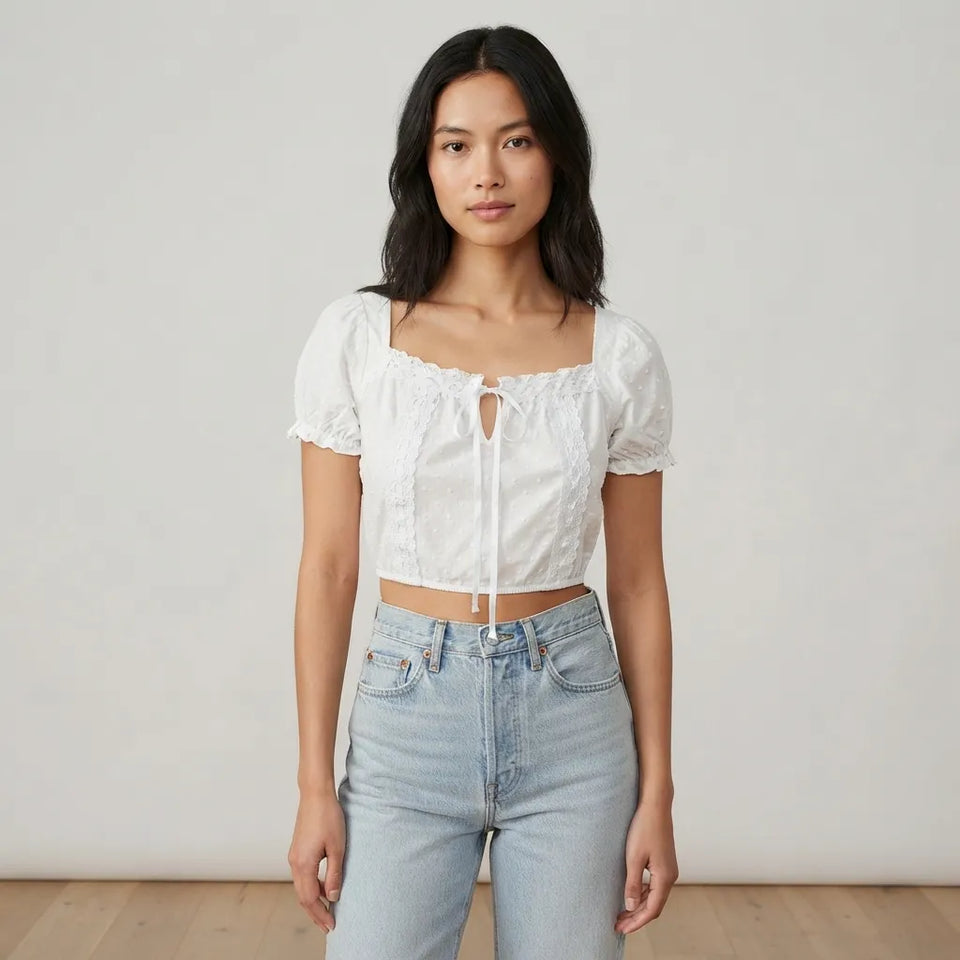 A woman modeling a white short-sleeve crop top with lace trim and a front tie closure, paired with light-wash blue jeans, against a plain off-white studio background.