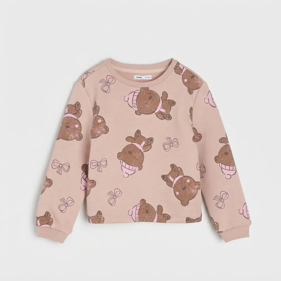 A children's long-sleeve crewneck sweatshirt in a dusty pink color, featuring an all-over print of teddy bears wearing winter hats and scarves, along with bow motifs, laid flat against a light gray background.