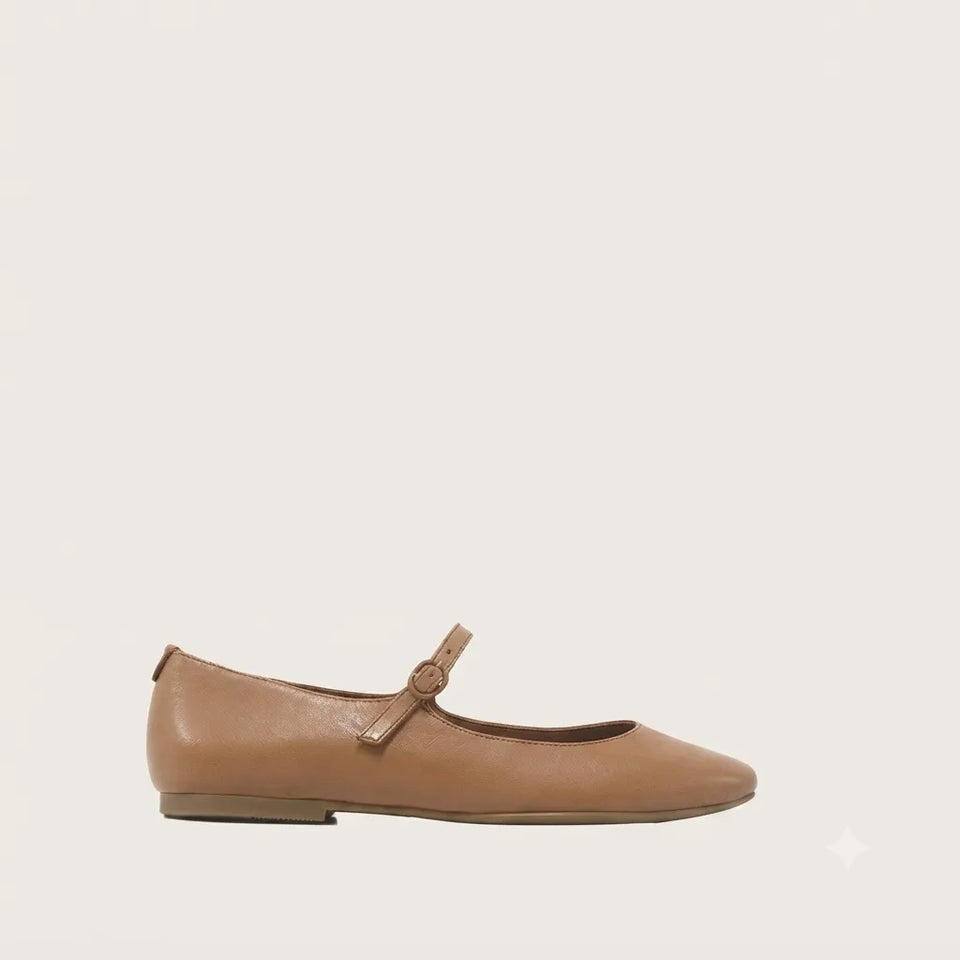 A light brown leather Mary Jane flat shoe with a small decorative strap and buckle detail across the instep, shown in profile against a solid off-white background.