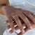 A close-up of a person's hands wearing a delicate gold ring with small engraved text, styled with a white top.
