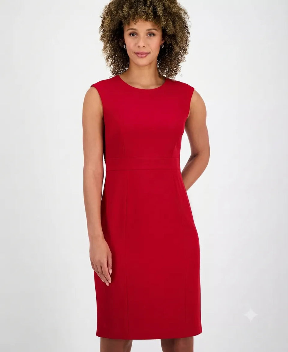 A woman with curly brown hair wearing a bright red sleeveless knee-length dress, standing against a light gray studio background.
