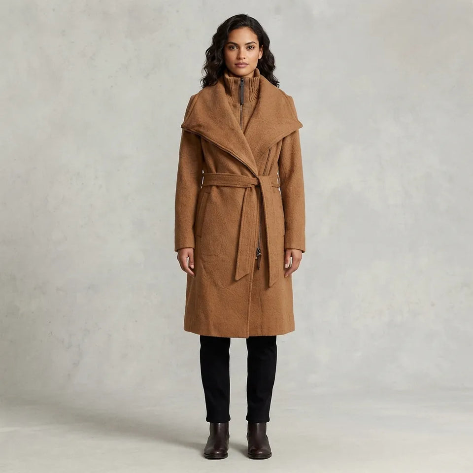 A woman modeling a camel brown wrap coat with a wide shawl collar and matching waist tie, paired with black pants and brown ankle boots, against a textured light gray studio backdrop.