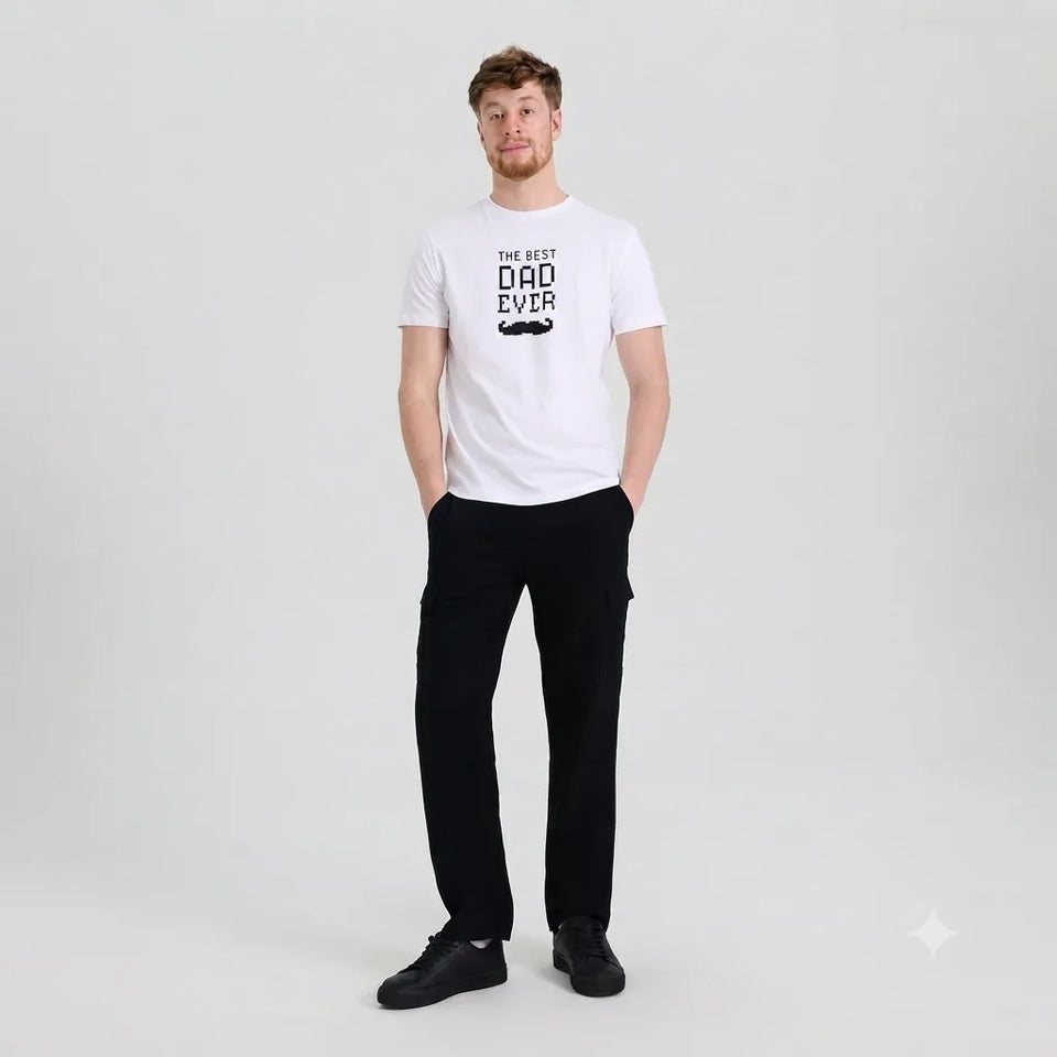 A man modeling a white crew-neck t-shirt with a black graphic print that reads 'THE BEST DAD EVER' with a mustache illustration, paired with black cargo pants and black sneakers, against a light gray studio background.