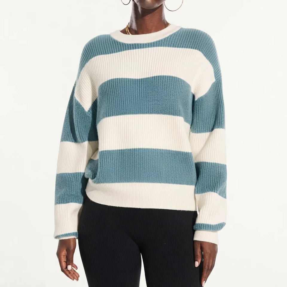 A woman wearing a ribbed knit sweater with wide horizontal stripes in teal blue and off-white, paired with black pants, against a plain white studio background.