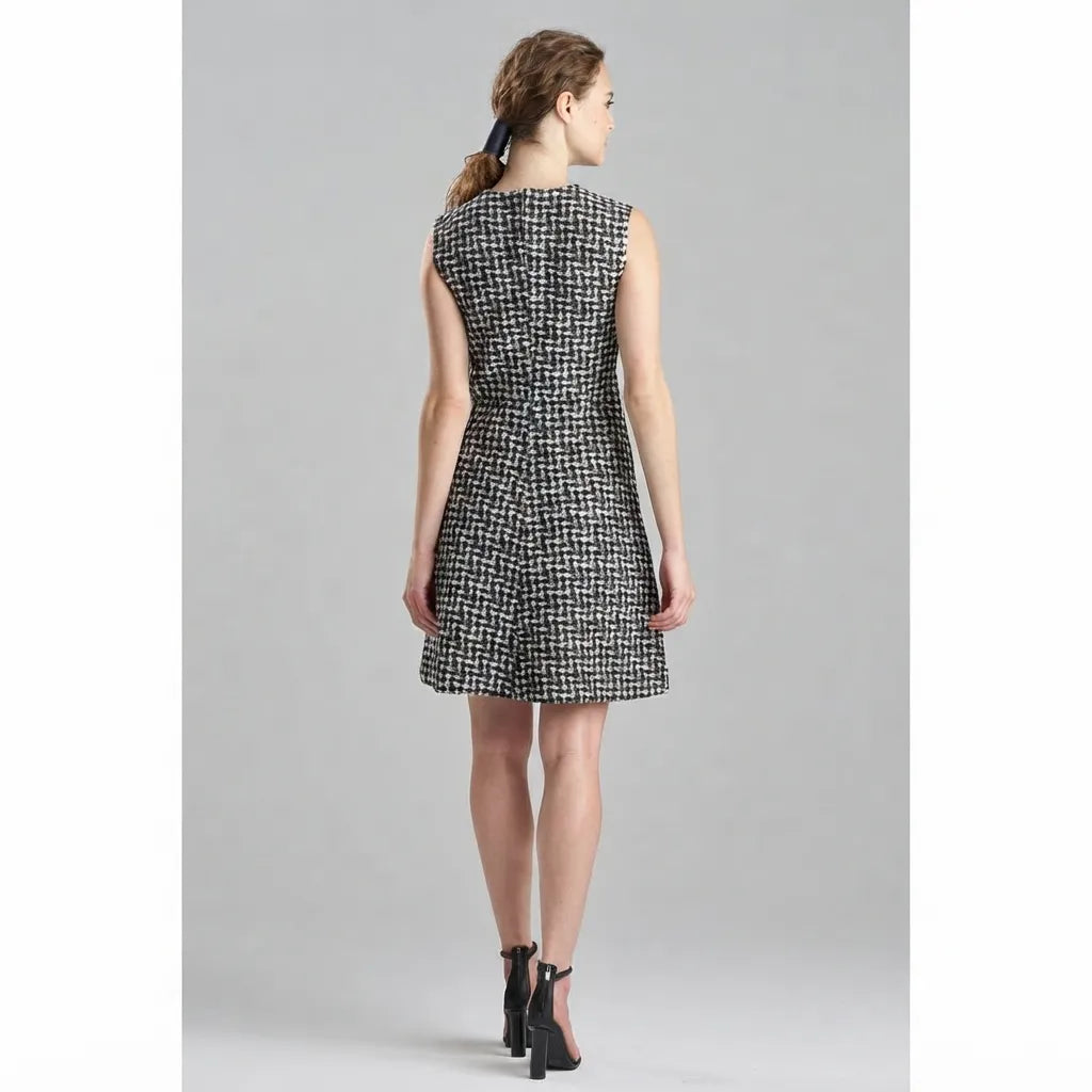 A woman viewed from the back wearing a sleeveless black and white micro-check tweed dress with a flared skirt, paired with black ankle-strap heels, against a solid gray studio background.