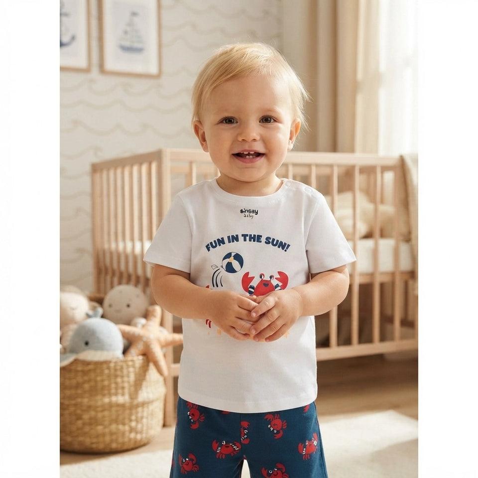 A smiling toddler stands in a softly lit nursery wearing a white short-sleeve T-shirt with a blue crab graphic and the words 'FUN IN THE SUN!' plus matching navy pants with red crab prints.