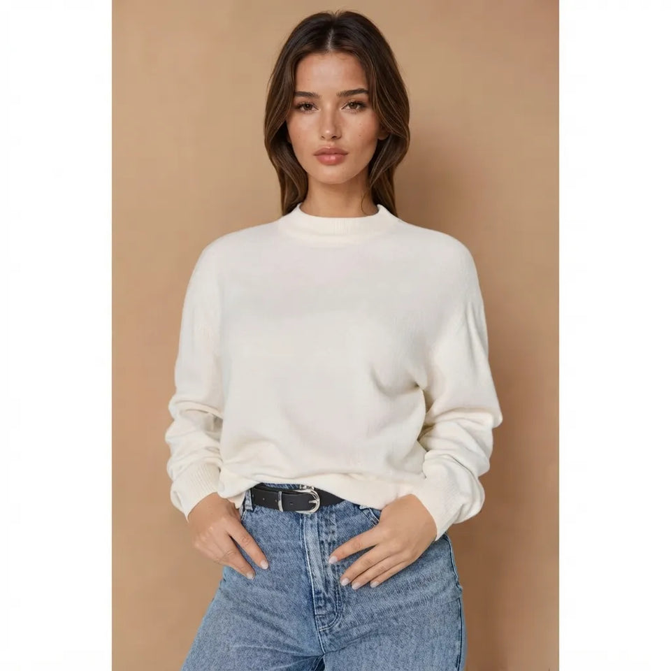 A woman modeling a cream-colored long-sleeve knit sweater with a mock neck, paired with blue jeans and a black belt, against a solid tan studio background.