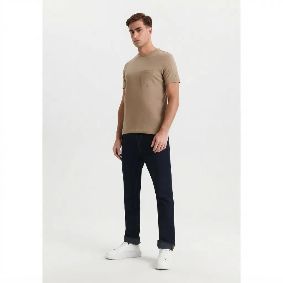 A man modeling a beige short-sleeve crewneck t-shirt with a chest pocket, paired with dark navy jeans and white sneakers, against a plain white studio background.