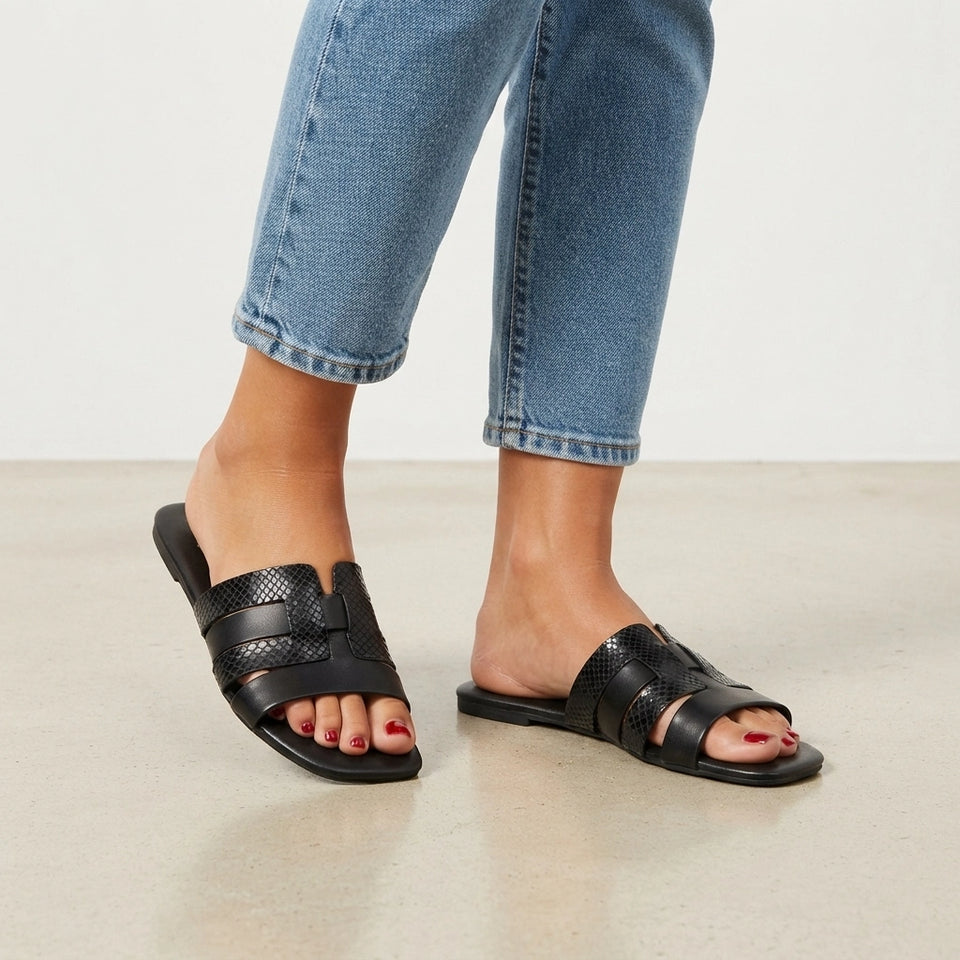 A person wearing black slide sandals with textured straps and blue cropped jeans, standing on a light beige floor against a white wall.