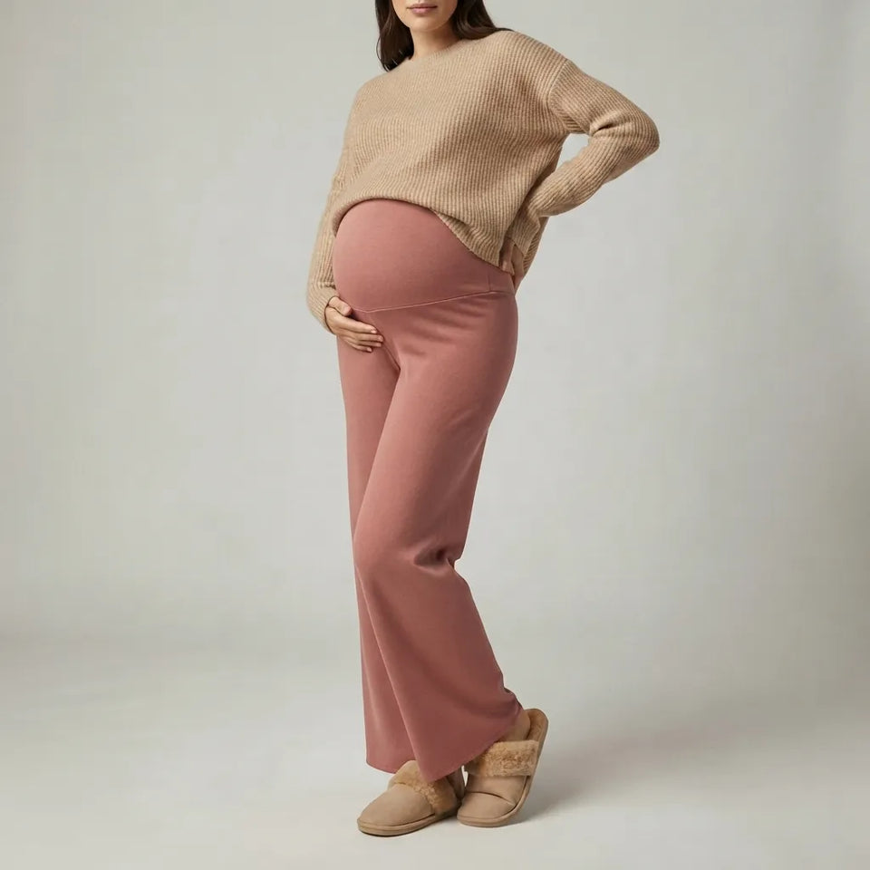 A pregnant woman wearing a beige ribbed knit sweater and dusty rose maternity pants, standing against a light gray studio background.