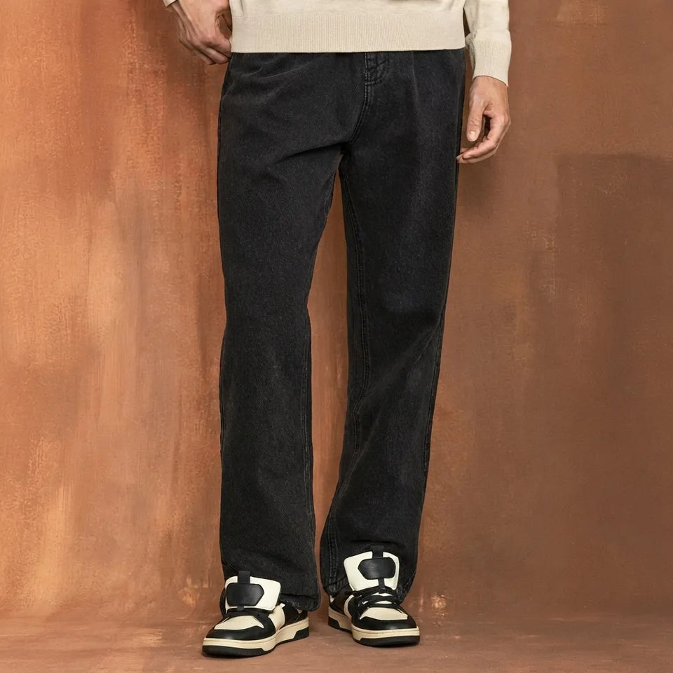 A person wearing dark charcoal gray wide-leg jeans, a cream knit sweater, and black-and-white sneakers, standing against a textured warm brown studio backdrop.