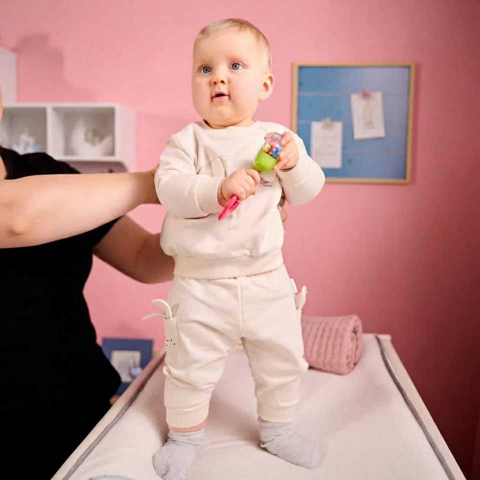 A baby wearing a cream-colored long-sleeve top and matching pants stands on a changing table while being supported by an adult. The baby is holding a small rattle in a pink room with a pink wall and a blue framed board in the background.
