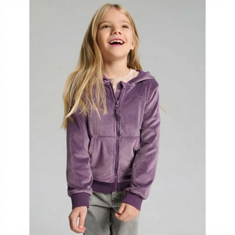A young girl with long blonde hair smiling and looking upward, wearing a purple zip-up hooded jacket with a plush velvety texture, paired with gray pants, against a solid light gray studio background.