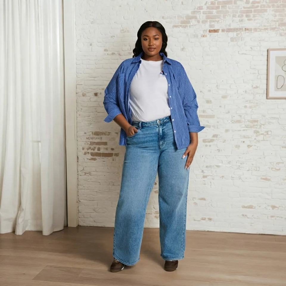 A woman modeling a pair of wide-leg light blue jeans, styled with a white t-shirt and a blue button-down shirt, standing in front of a white brick wall.