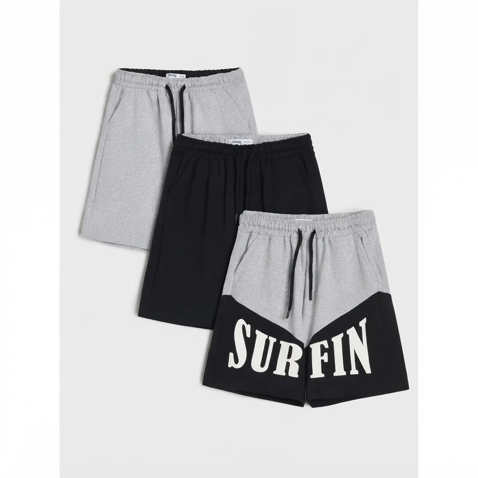 Three pairs of women's colorblock athletic shorts are displayed in a stacked arrangement on a light gray background. The set includes heather gray and black shorts, with the front pair featuring a colorblock design with 'SURFIN' printed in white block letters across the lower half.