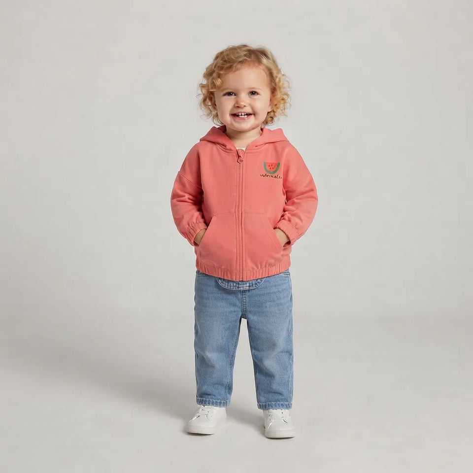A toddler with curly blonde hair stands against a light gray studio background wearing a coral pink zip-up hooded jacket, blue jeans, and white sneakers. The jacket has a small embroidered logo on the left chest.