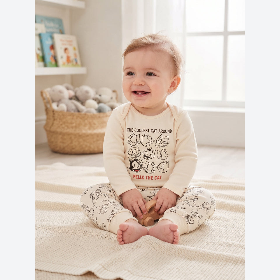 A baby sitting on a cream knit blanket in a bright room, wearing a cream long-sleeve top with a black cat graphic and text reading 'THE COOLEST CAT AROUND FELIX THE CAT,' paired with matching cream patterned pants.