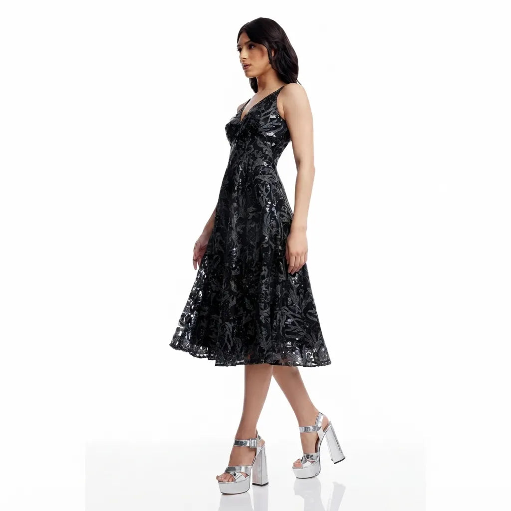 A woman modeling a sleeveless black midi dress with a textured, slightly shiny floral or abstract pattern, paired with chunky silver platform heels, against a plain white studio background.