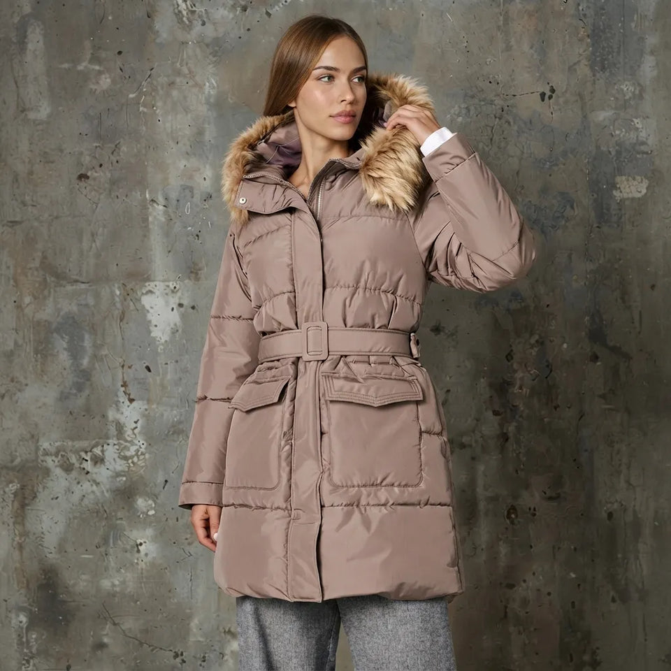 A woman wearing a taupe-brown puffer coat with a faux-fur trimmed hood and a matching belt, standing against a textured gray concrete wall.