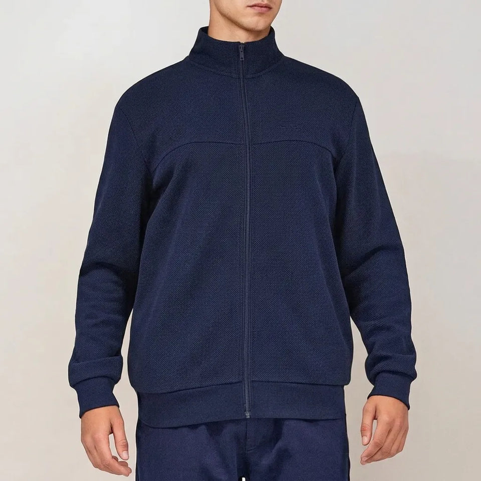 A man wearing a navy blue full-zip fleece jacket with a mock neck collar, paired with matching navy pants, against a plain light beige background.
