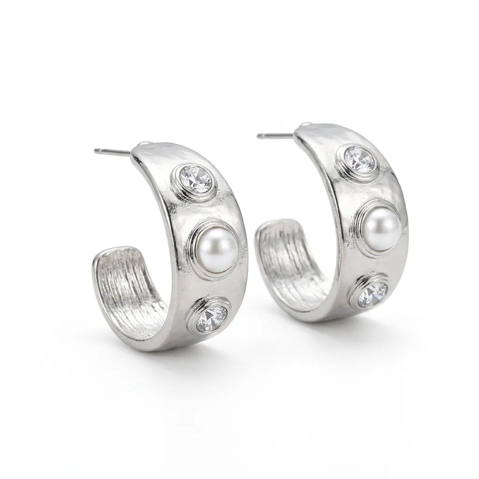 A pair of silver crescent-shaped hoop earrings decorated with three white faux pearls and three clear rhinestones on each earring, set against a plain white background.