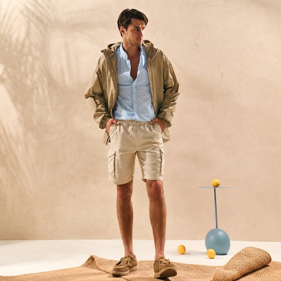 A man stands in a studio wearing a light brown jacket over a light blue button-down shirt and beige cargo shorts, paired with tan sneakers. The background is a warm beige textured wall with soft shadows, and a small blue decorative stand with yellow spheres sits to the side.