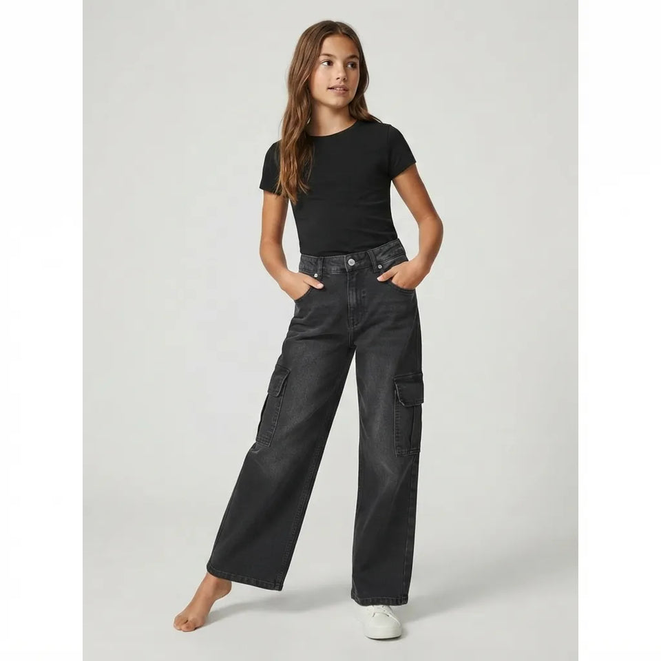 A young girl modeling a pair of dark gray wide-leg cargo jeans with a black t-shirt and white sneakers, standing against a light gray studio background.