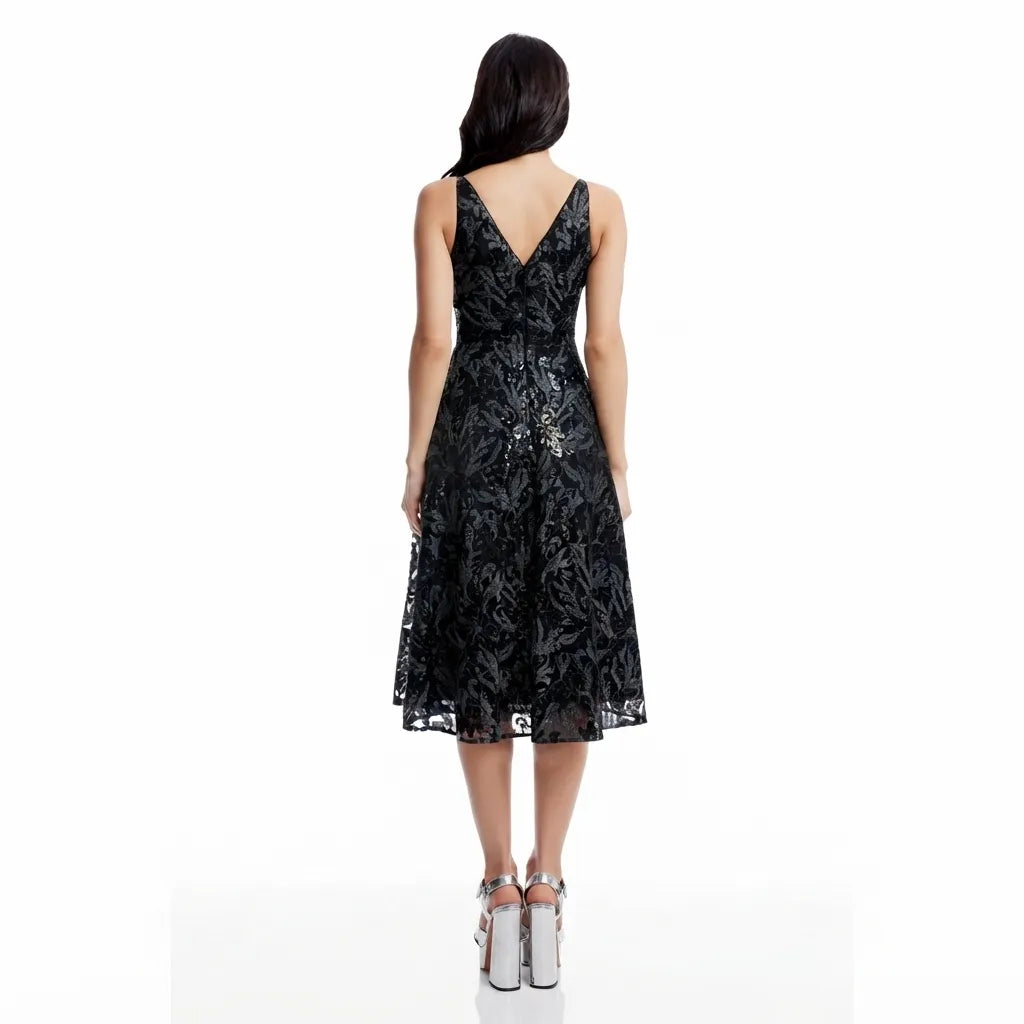 A woman viewed from the back wearing a sleeveless black lace midi dress with a V-neckline and a flared skirt, paired with metallic high heels, against a plain white studio background.
