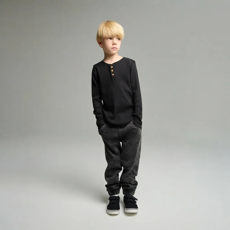 A young boy with blonde hair stands against a neutral gray studio background wearing a black long-sleeve henley shirt with three gold buttons, dark gray textured jogger pants, and black sneakers.