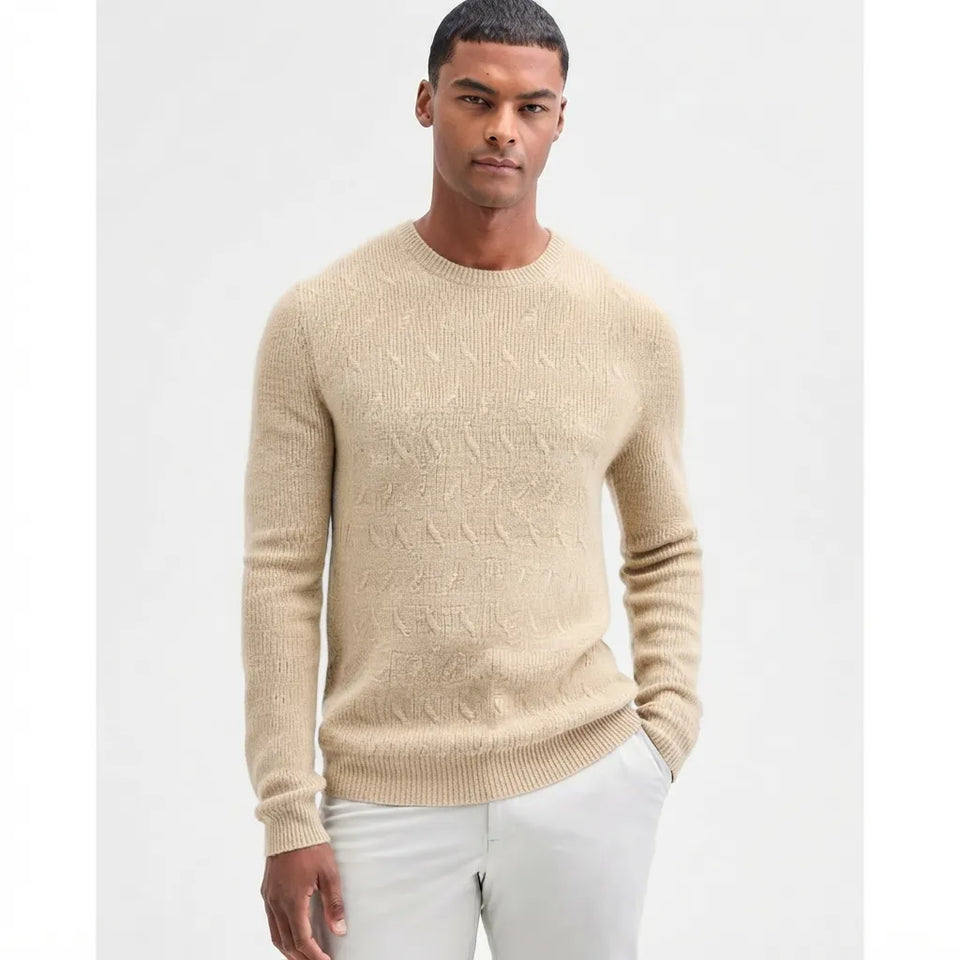 A man wearing a beige textured knit crewneck sweater and white trousers, standing against a plain light gray studio background.