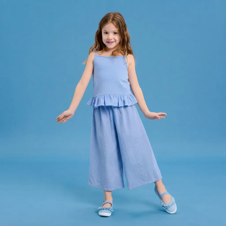 A young girl modeling a light blue two-piece outfit consisting of a sleeveless top with a ruffled hem and matching wide-leg pants, set against a solid blue studio background.