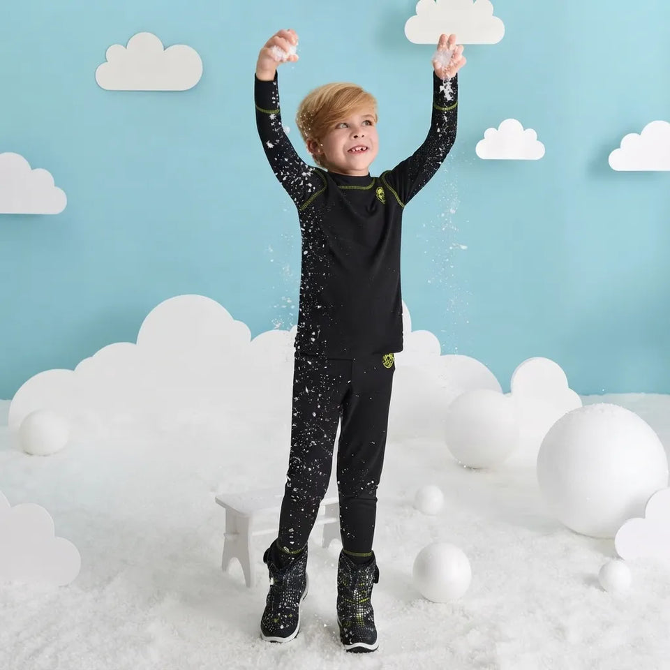 A young boy wearing a black full-body snowsuit with small white speckles and lime green accents stands in a snowy play area with a light blue wall decorated with white clouds.