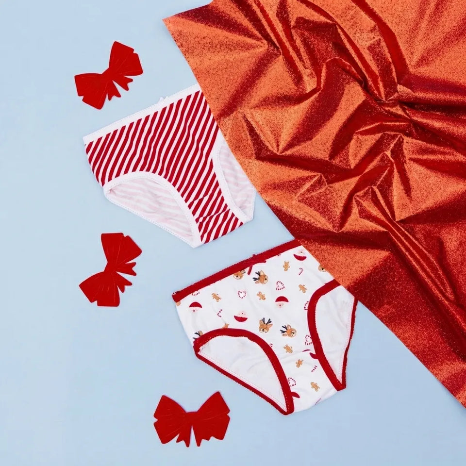 Two pairs of women's briefs laid flat on a light blue background with decorative red bows and a large metallic red fabric sheet. The top pair has thin red and white stripes, and the bottom pair is white with small holiday-themed prints and red trim.