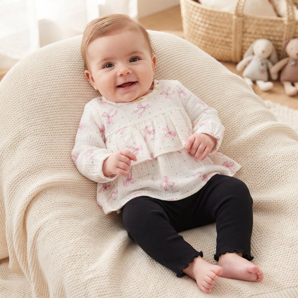 A baby sitting on a cream knit beanbag chair, wearing a white long-sleeve ruffle top with pink bow prints and black pants.