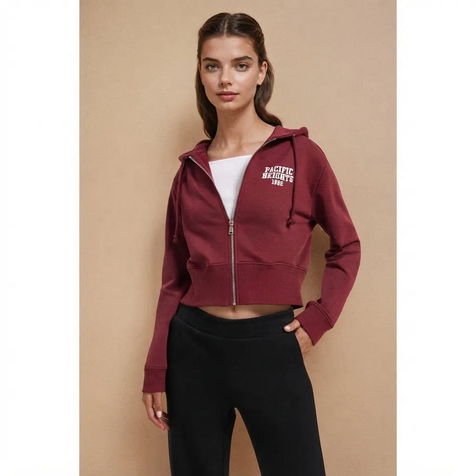 A woman modeling a cropped zip-up hoodie in burgundy with white 'PACIFIC BAYOUTED' text on the left chest, worn over a white top and black pants against a beige studio background.