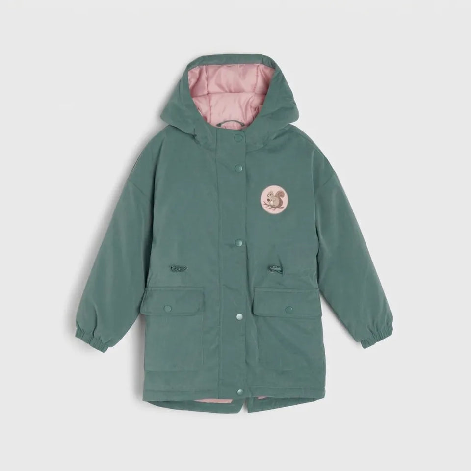 A children's hooded jacket in muted sage green with a pink quilted lining inside the hood, featuring a circular embroidered patch on the left chest and snap-button front closure, shown against a light gray background.