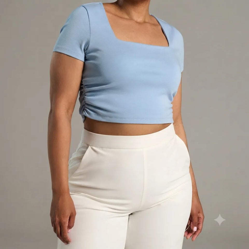 A model wearing a light blue short-sleeve crop top with a square neckline and white high-waisted pants, shown from the neck down against a solid gray studio background.