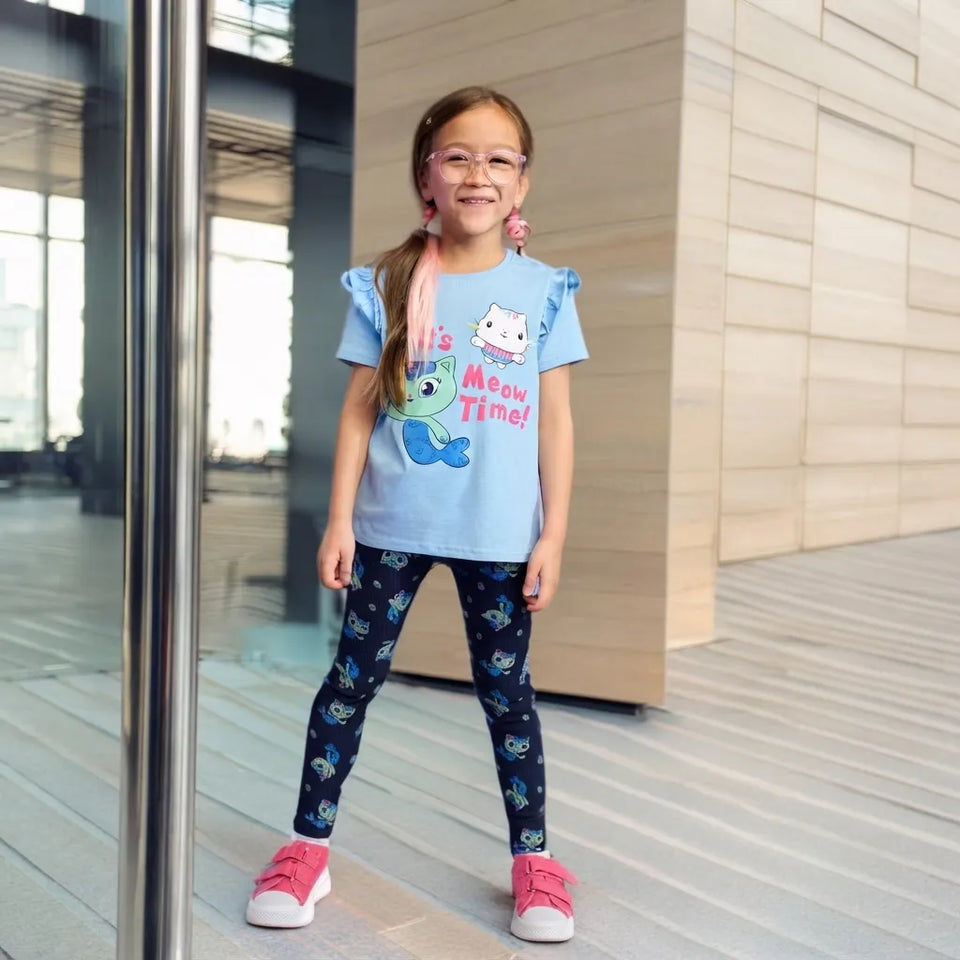 A young girl with glasses and a ponytail stands in front of a modern glass building, wearing a light blue graphic t-shirt with a cartoon character and the text 'Meow Time!', dark patterned leggings, and pink sneakers.