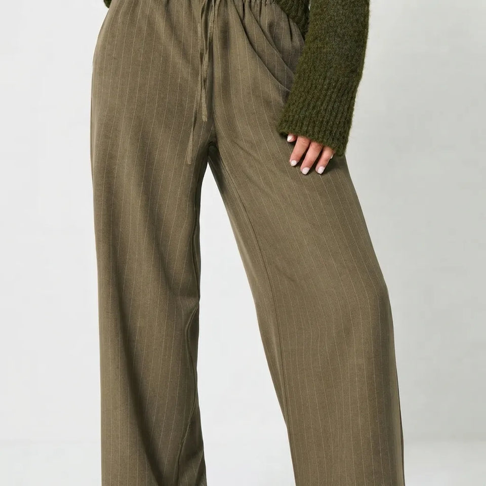 A close-up of a person wearing olive green wide-leg pinstripe trousers with a drawstring waist, styled with an olive green knit sweater against a light gray studio background.