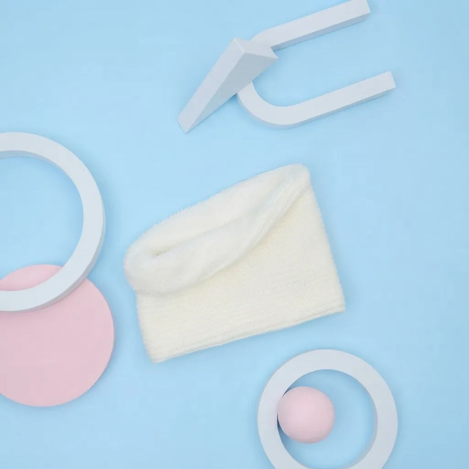 A folded cream-colored plush fabric item, possibly a pouch or small bag, styled in a minimalist flat-lay with white geometric rings, a pink circle, and a white arrow-shaped object on a light blue background.
