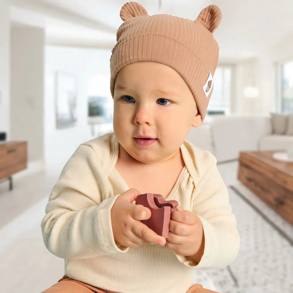 A baby wearing a beige ribbed knit beanie with bear ears and a cream ribbed long-sleeve top, holding a small dusty rose gift box toy in a bright, modern living room.