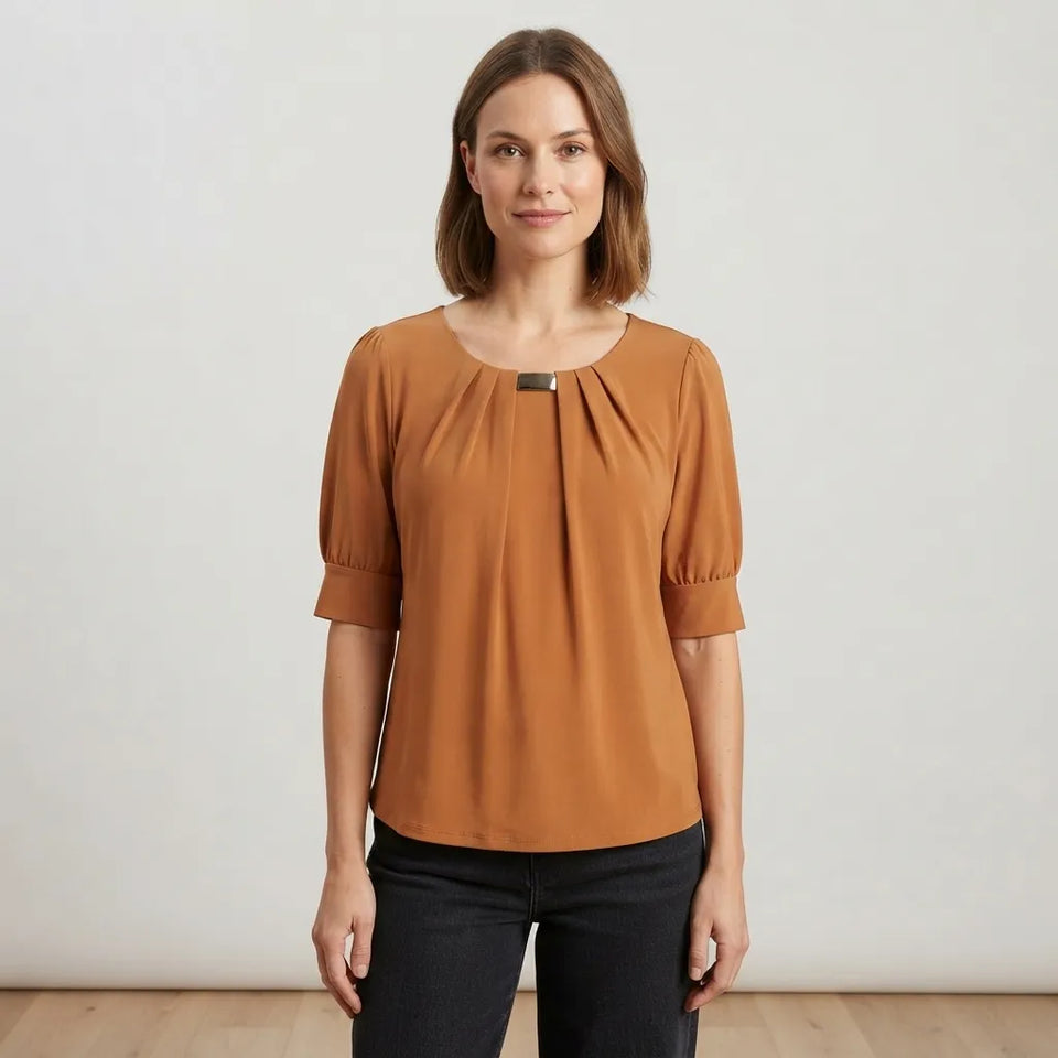A woman modeling a rust-colored short-sleeve blouse with a gathered neckline and a small metallic accent, paired with black pants, against a neutral studio background.