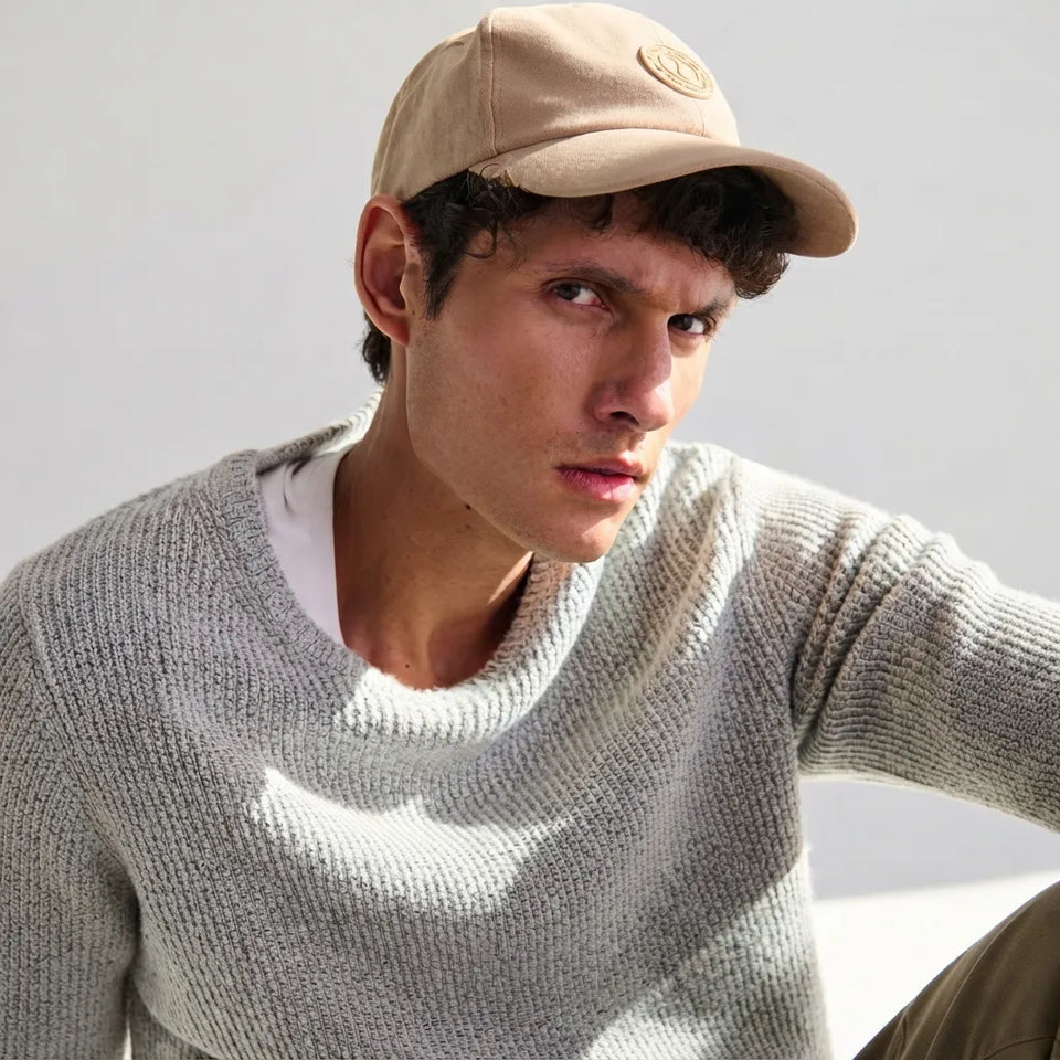 A man wearing a beige baseball cap with a circular logo patch and a light gray ribbed knit sweater, photographed against a clean white background.