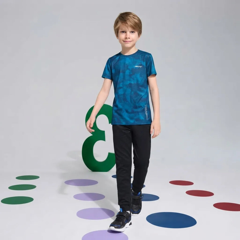 A young boy wearing a blue camouflage short-sleeve athletic t-shirt, black pants, and black sneakers, walking on a light gray studio floor decorated with colorful circular shapes.